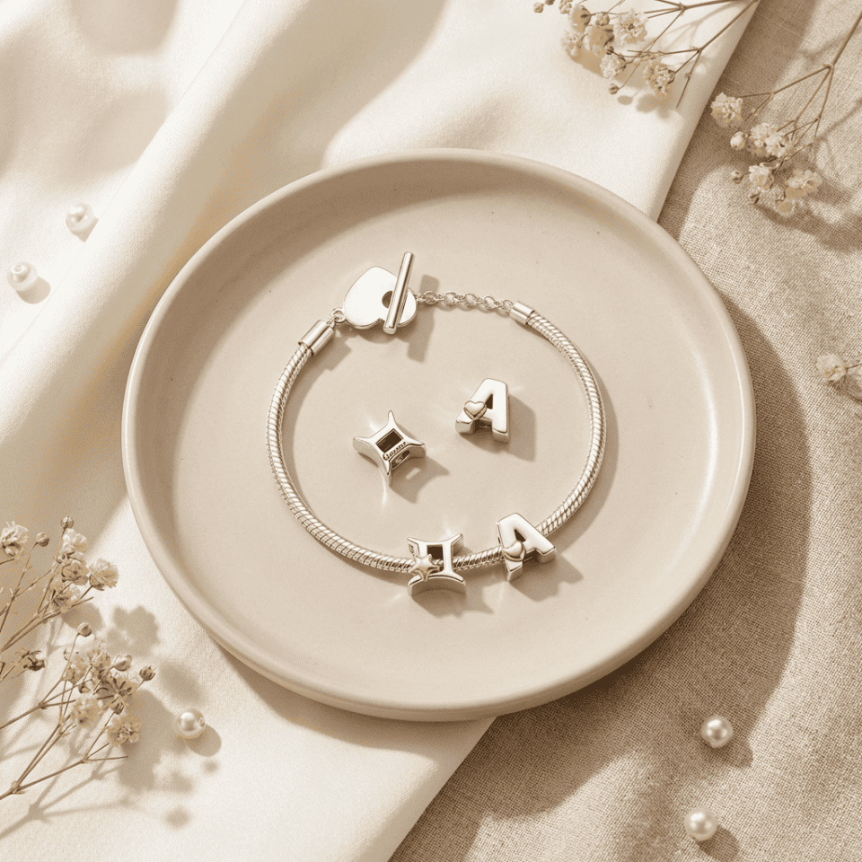Bracciale elegante con charm a forma di lettere, in argento, su piatto di ceramica beige, decorato con fiori secchi e perle, ideale come regalo di gioielli raffinati e di tendenza.